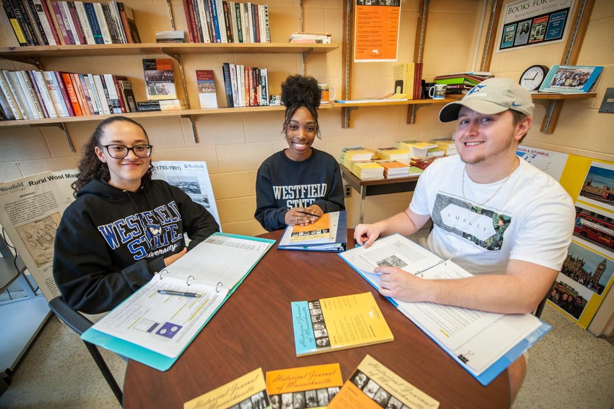 Three history students sitting at a table, smiling, surrounded by bookshelves filled with books.