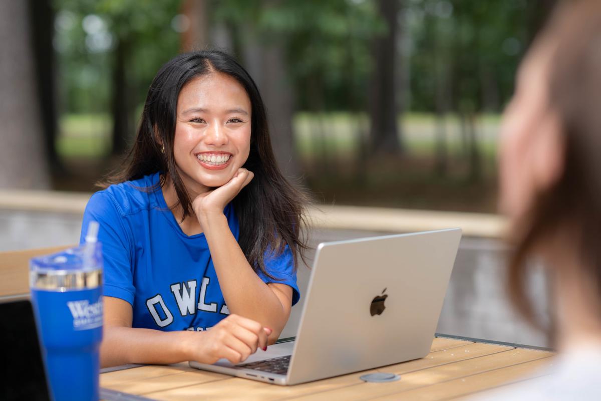 Student studying outside with a laptop while talking to another student.