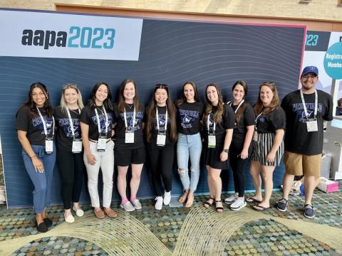 A photo of Andrew Allard, Nicole Clemente, Joelle Encarnacion, Melinda Hurteau, Giselle Lemus Tejada,  Kadeja Miller, Jennifer Petrucci, Taylor Saimeri, Mark Sigwart, and Hailey Whipple (not in order), smiling in front of a dark-blue backdrop AAPA 2023 poster.