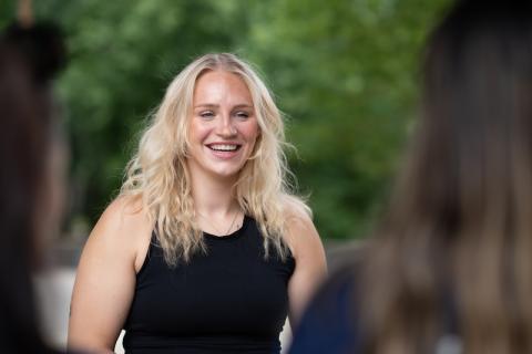 Jessica Gardner, a blond young woman in a black tanktop, smiles at a group of students blurred and in the corner of the left-hand side of the photo.