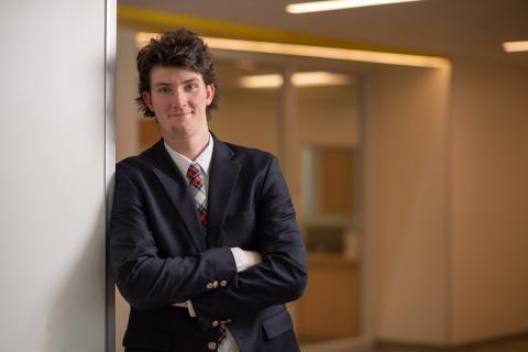 Seamus Mitchell, class of 2024 and a double-major in criminal justice and English, is dressed in a black suit coat and red tie. He is smiling and leaning against a white wall, with a golden interior behind him. Both arms are folded across his chest.