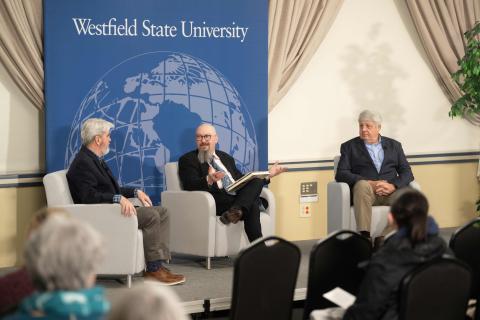 Three men sit in chairs on a stage in front of an audience