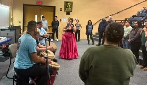 Woman sings to audience with row of drummers and dancers