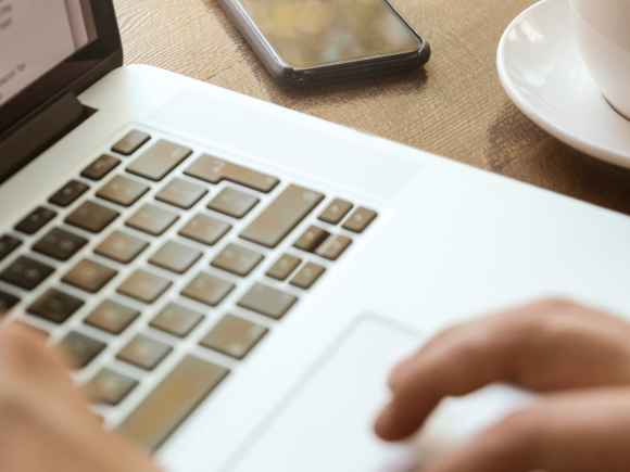 Close up of hands on the keyboard of a laptop, there is a smartphone and cup of coffee next to the laptop