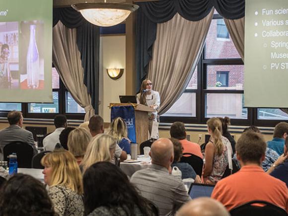 A crowd listens to a presenter in a conference-like setting.