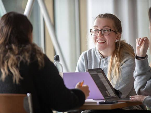 Three students smiling while engaging in conversation while working on laptops.