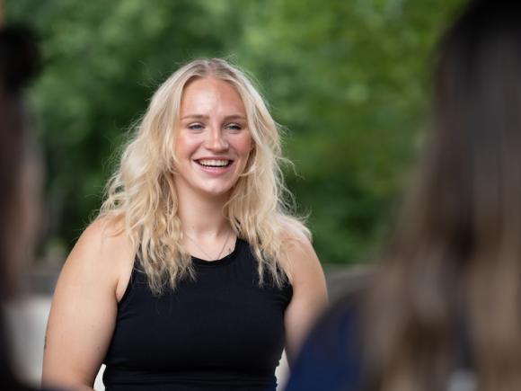 Jessica Gardner, a blond young woman in a black tanktop, smiles at a group of students blurred and in the corner of the left-hand side of the photo.