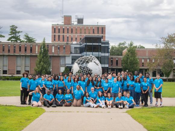 A photo of the Urban Education Summer Bridge Program students wearing a turquoise-colored T-shirt and shorts. They pose in front of the campus globe, which features the brick Ely building in the background.