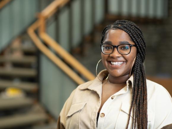 Sydney Maiden, a young, African American woman dressed in a beige, button-down short sleeve shirt. She also wears black glasses and hoop earrings. Her hair is swept into a side ponytail.