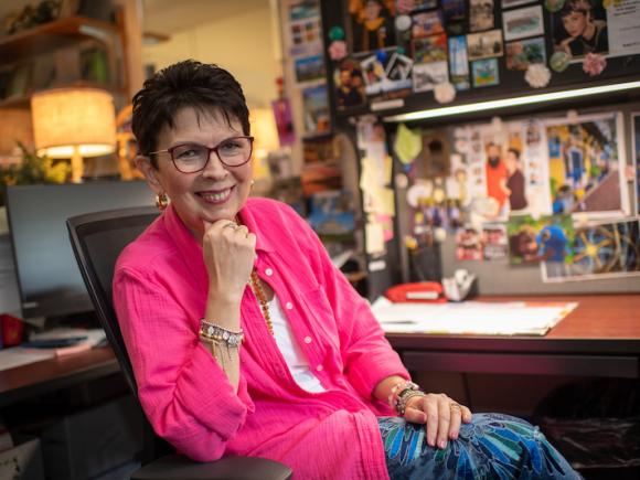 Regina, wearing a pink jacket, bracelets, earrings, glasses, and blue pants, is sitting in her office with her hand tucked under her chin and smiling.