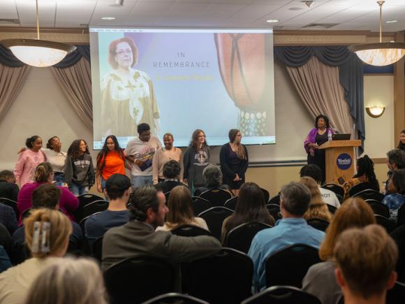 A crowd full of people sit in front of a presentation titled: The Voice of Resilience. One of the presenters stands in front of a podium and addresses the crowd.