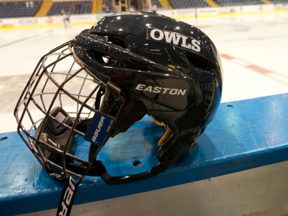 A close-up shot of a navy blue hockey helmet that has "Owls" printed in white letters on its side. It's resting on the edge of the wall of a hockey rink.