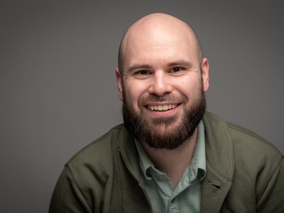 Man smiling wearing a green coat, green shirt and against a grey background.