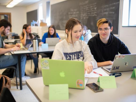 Two students with laptops smiling in math class. One student is wearing a WSU sweatshirt the other a black sweatshirt.