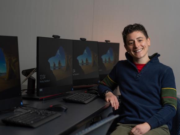 Ab Mosca, sitting in front of a row of black computers. They are wearing a long sleeve, blue sweatshirt and have one arm resting on the desk beside them.