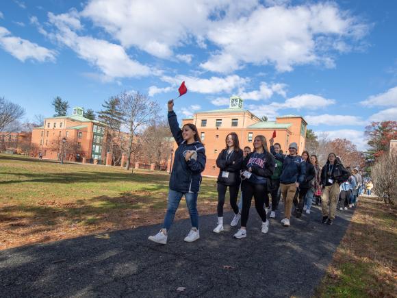 Campus tour with tour guide holding flag with line of students following. Blue sky and campus building behind them.