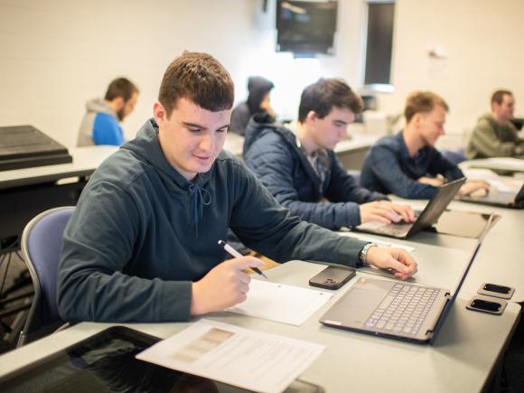 Computer science student writing in front of laptop wearing blue long sleeve shirt.