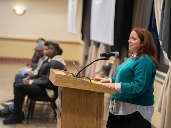 Rebecca Ashley, teacher at Canton High School, dressed in a long-sleeve, green sweater and speaking at a wooden podium as part of the The Impact of Learning about Social Class in Twelfth Grade ELA event on campus.