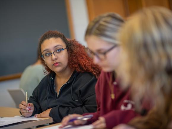 Three students in history class writing with one student looking up with long red hair and black shirt.