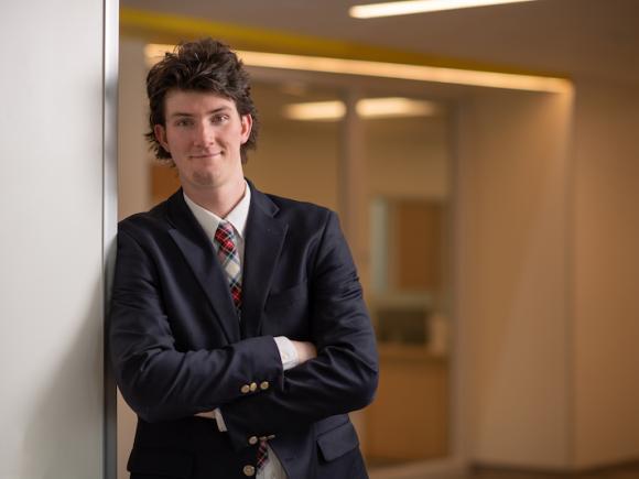 Seamus Mitchell, class of 2024 and a double-major in criminal justice and English, is dressed in a black suit coat and red tie. He is smiling and leaning against a white wall, with a golden interior behind him. Both arms are folded across his chest.