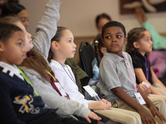 Group of five children sit in a row listening to a speaker while one child raises a hand