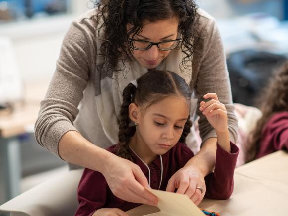 Female teacher stands behind a young girl who sits at a table, leaning over a reading and writing project