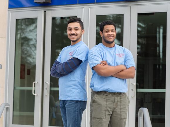 Two students in front of Parenzo wearing blue WSU shirts back to back with arms crossed.