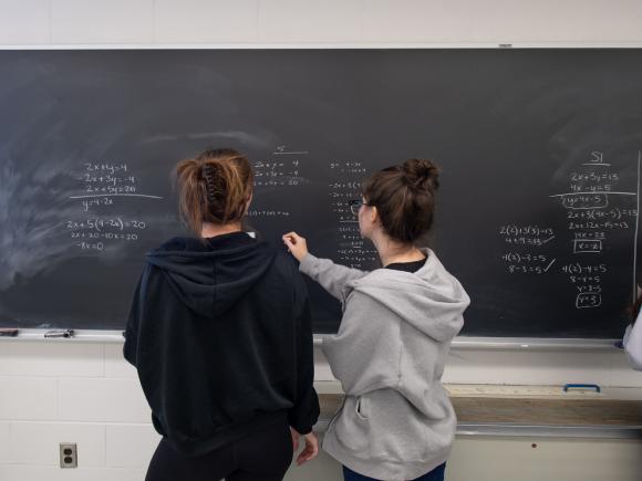 Two students with pony tails and hooded sweatshirts doing math equations on chalkboard