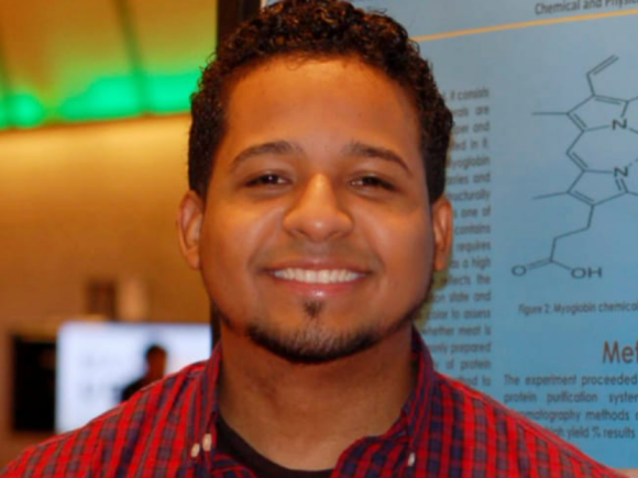 Chemistry alumni smiling wearing red and black plaid shirt in front of CURCA poster.