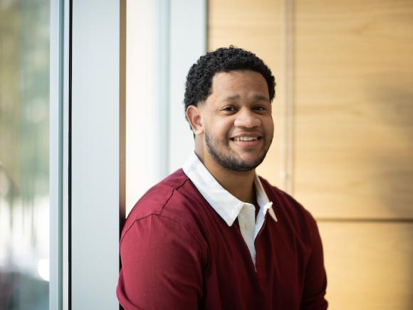 Carlos Lantigua Bari, class of 2025, is dressed in a red, long-sleeved shirt with a white collar. He's sitting near a window with wood paneling behind him. He's smiling at the camera.