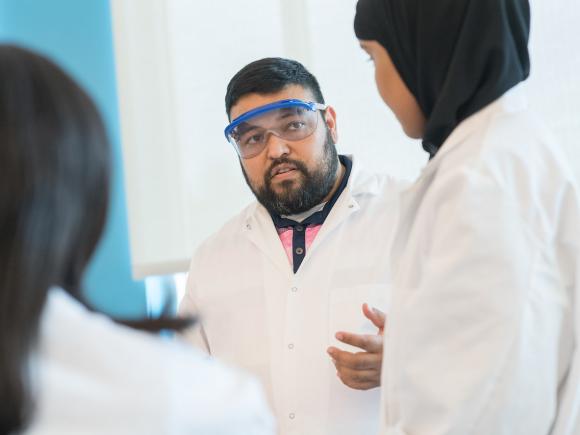 Chemistry faculty member wearing eye protection and white gown in a lab with two students.
