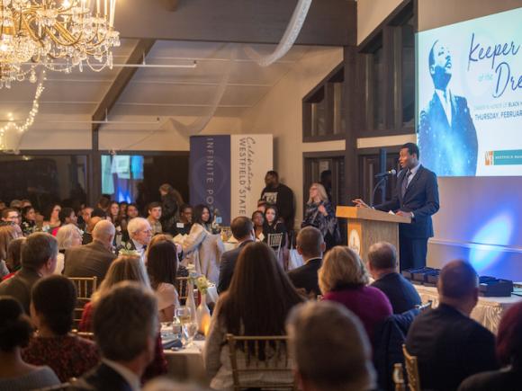 A photo of the Inaugural Keeper of the Dream dinner. Various tables with people sitting at them face a man speaking at a podium at the front of the room, and he stands in front of a projector screen depicting Martin Luther King Jr.