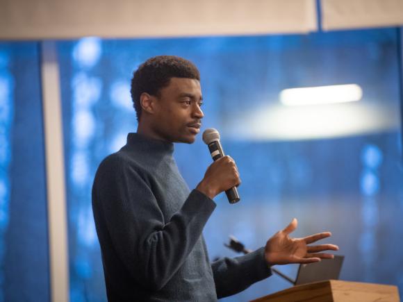 Zul Manzi, a Tanzanian-American content creator, is standing in front of a blue poster of trees. He is dressed in a long-sleeve, blue sweater and holds a microphone in his hand. He's speaking to a crowd of people who've gathered to watch his original show, "The Matimbulas".