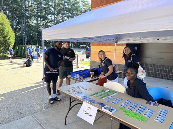 A white tent outside next to University Hall. Residential Assistants sit at a table under the tent and hands out prizes and goodies to RAs who attend the celebration. Two students stand in front of the tent, while there students sit inside of it.