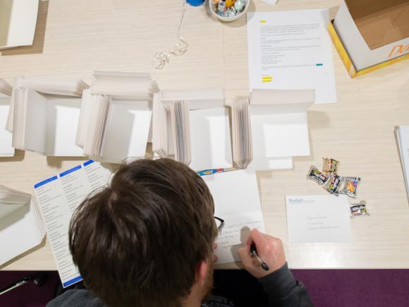 A close-up photo of a brown-haired student sitting at a white desk. He is writing hand-written thank-you cards as part of the Give a Hoot campaign. The table has stacks of cards, paper, and containers. He is writing a card, and has a pen in his hand. The angle is peeking over his shoulder with the photographer standing behind him.