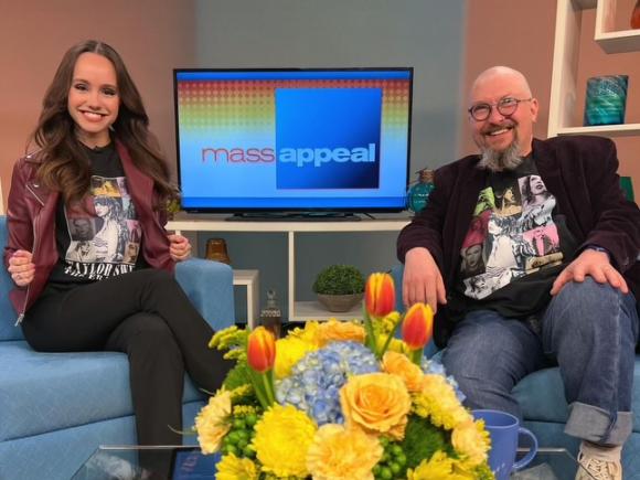 Dr. Christopher Cullen of Westfield State and Kayla Hevey of Mass Appeal sit in blue chairs next to each other. A yellow centerpiece of flowers sit in between them. Behind is a TV screen with the words "mass appeal" on it in a yellow, red, and white gradient backdrop. Cullen is dressed in jeans, a black overcoat, and a black undershirt. Heavy is in black trousers, a maroon overcoat, and an identical black undershirt.