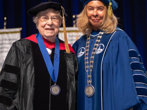 President Thompson and Dr. Catherine B. Shannon. President Thompson wears a blue robe, with Shannon wearing a black robe. They're smiling at the camera as Dr. Shannon is awarded the President’s Medal of Achievement for her contributions to the University.