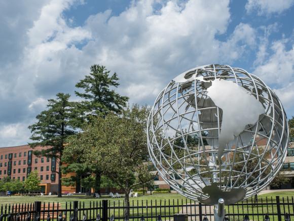 A photo of the campus globe during the summer season. The sky above is blue with clouds, and University Hall can be seen behind the globe itself.