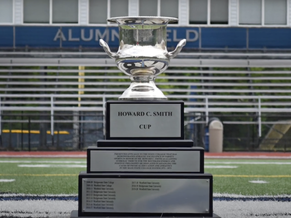 A close-up photo of the Howard C. Smith Cup trophy. It's a large silver chalice with three tiers of printing on its lower levels. It sits on the football field at the University.