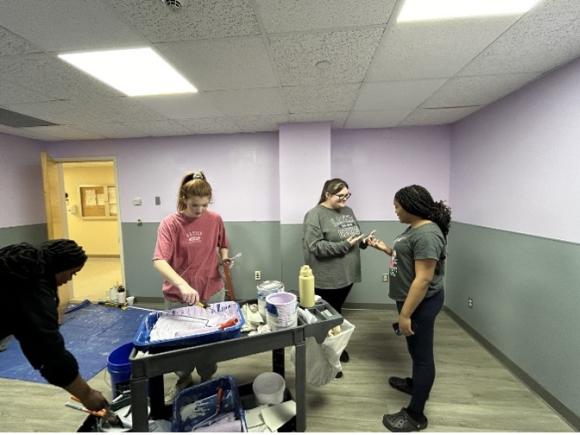 Students creating a pediatric waiting room at MiraVista Behavioral Health Center in Holyoke, MA, as part of the nursing department's the Psychiatric Nursing class. Four students are painting a white room to green, and use paints provided by a small, portable, black worktable.