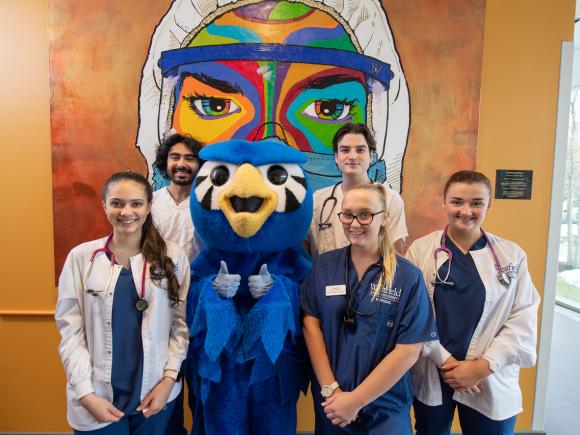 Nursing students smiling in front of a nursing mural with Nestor giving thumbs up.