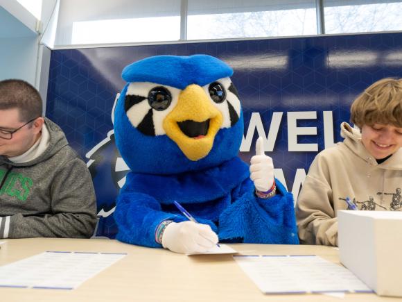 Blue owl mascot shows a thumbs up while two students write notecards