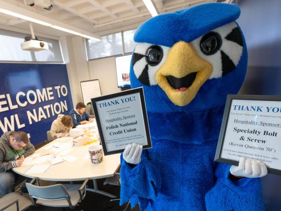 Blue owl mascot holds up signs while people write in the background