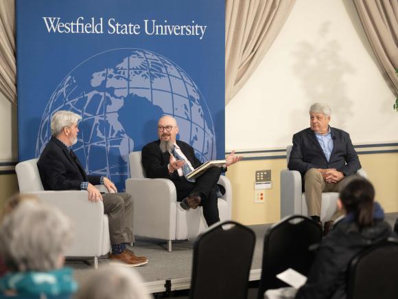 Three men sit in chairs on a stage in front of an audience