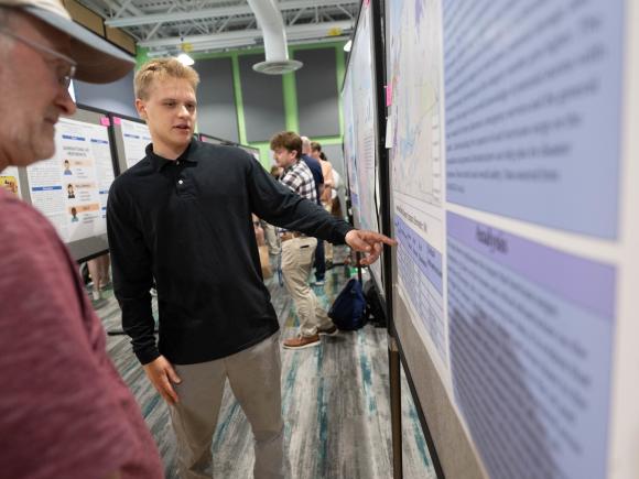 Student presenting research by pointing to a poster during an academic event.