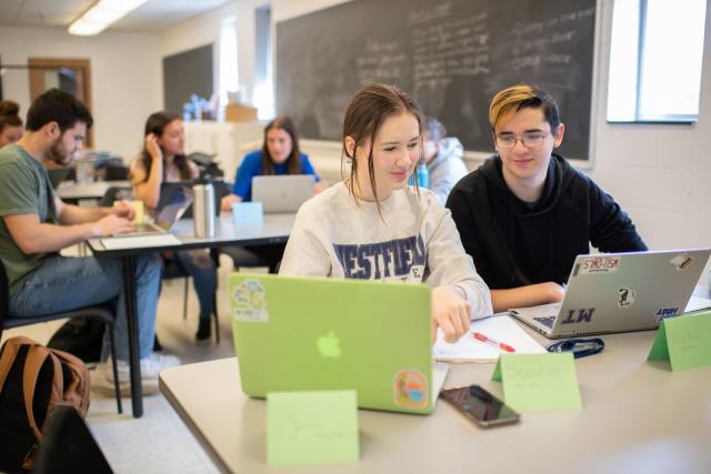 Two students with laptops smiling in math class. One student is wearing a WSU sweatshirt the other a black sweatshirt.