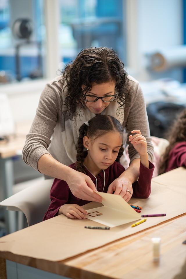 Education student assisting an elementary student with a project. Crayons on desk with paper.