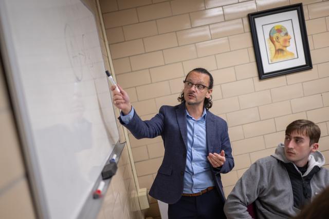 Faculty member teaching at the front of a classroom, pointing to information on a whiteboard.