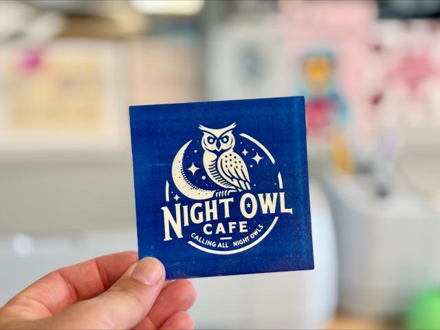 hand holding a sticker labeled "night owl cafe", blue background