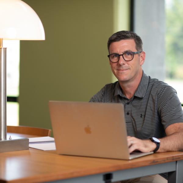 Math student wearing striped polo shirt and glasses sitting at a desk with Mac computer